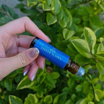 Een hand houdt een essentiële olieroller vast. Op de olieroller zit een donkerblauw label waarop staat: 'lekker slapen'. Op de achtergrond zijn groene bladeren.