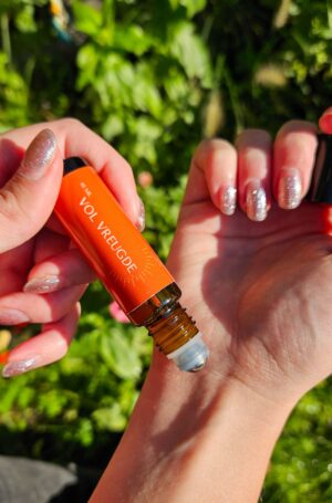 Een essentiële olie roller met een oranje label waarop staat: Vol vreugde. Je ziet twee handen. De ene hand rol met de olieroller op de pols van de andere hand. Op de achtergrond zijn groene bladeren te zien.