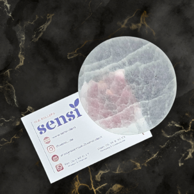 Een witte ronde schijf van een seleniet oplaadsteen. Deze ligt op een visitekaartje van Sensi-olie.