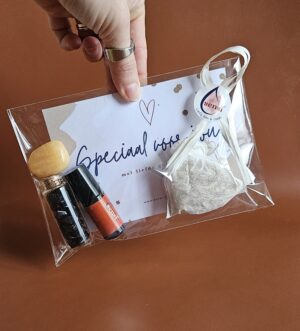 Een hand die een pakketje van transparant folie vasthoudt voor een bruine achtergrond. In het pakket zit een calciet trommelsteen, een wensbuisje met blauwkwarts steentje, een vol vreugde essentiële olie roller en een zakje ginseng thee. Ook zit er een wit kaartje in waarop staat: 'speciaal voor jou, met liefde gemaakt'. Op het kaartje staan ook een gouden hartje en wat gouden spetters. Op het pakketje zit een sticker met een wit lint. Op de sticker staan Sensi liefs Kim & Daan.
