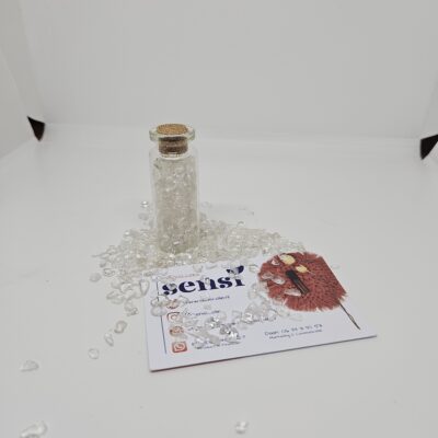 Een glazen buisje met transparante bergkristal erin. Ook om het flesje heen liggen stukjes bergkristal. Ervoor ligt een visitekaartje van Sensi-olie. De achtergrond is wit.